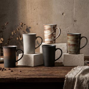 Può includere: Una collezione di sei tazze in ceramica in vari colori e texture. Le tazze sono esposte su una superficie di legno e blocchi di cemento. Le tazze hanno un design minimalista con manico e forma cilindrica.