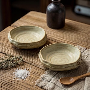 Puede incluir: Dos cuencos de cerámica hechos a mano, de color claro, con un aspecto rústico y texturizado. Los cuencos están sobre una superficie de madera, junto a una pequeña botella oscura, sal y una cuchara de madera. Los cuencos tienen un diseño poco profundo y redondo.