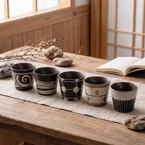 May include: Five small, dark brown ceramic cups with varying white patterns. The cups are arranged on a beige runner atop a wooden table, with a book and natural decor in the background. Each cup has a unique design.