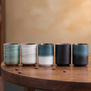 Może przedstawiać: Pięć cylindrycznych ceramicznych kubków w różnych kolorach, w tym turkusowym, białym i czarnym, z brązowymi wnętrzami, ustawionych na drewnianym stole. Ziarna kawy są rozrzucone wokół kubków.