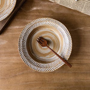 May include: A ceramic plate with a textured, cream-colored rim and a brown, spiral-patterned center. A wooden fork rests on the plate. The plate is on a wooden surface, suggesting a rustic or handmade aesthetic.