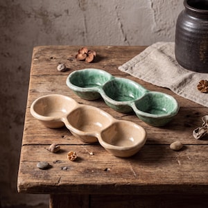 May include: Two elongated ceramic serving dishes, each featuring three connected circular bowls. One dish is finished in a glossy sage green glaze, while the other is finished in a glossy cream glaze. Both items have irregular, organic edges.