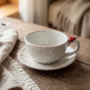 May include: A handmade ceramic teacup and saucer set. The cup is off-white with a textured surface and a red heart-shaped accent on the handle. The saucer is a matching off-white color. The set is placed on a rustic wooden surface.