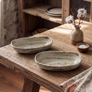 Op de afbeelding: Twee ovale keramische serveerschalen met een rustiek, getextureerd ontwerp. De schalen zijn lichtbeige met bruine strepen en spikkels, op een houten tafel. Een kleine vaas met katoenen stelen en een houten kom met zout staan ook op de tafel.