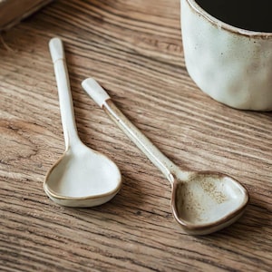 May include: Two ceramic spoons in cream with a light brown rim. The spoons have long, slender handles with a white tip. The spoons are on a wooden surface, with a ceramic mug in the background.