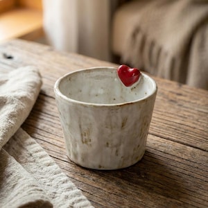 Könnte beinhalten: Eine kleine, cremefarbene Keramikschale mit strukturierter Oberfläche und einem glänzenden roten Herz am Rand. Die Schale steht auf einer rustikalen Holzoberfläche, mit einem neutralfarbenen Tuch im Vordergrund.