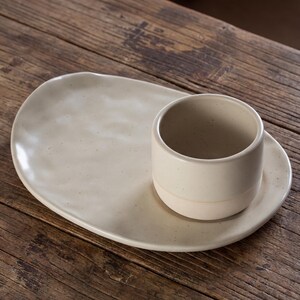 Minimalist handmade ceramic cup and oval saucer set on rustic wooden table, speckled stoneware finish