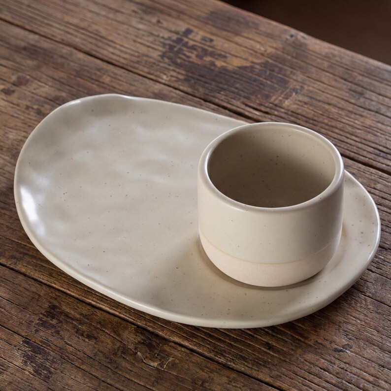 Minimalist handmade ceramic cup and oval saucer set on rustic wooden table, speckled stoneware finish