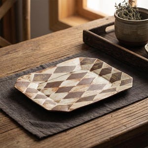 Op de afbeelding: Een rechthoekige keramische serveerschaal met een ruitpatroon in bruin- en crèmetinten. De schaal staat op een donkergrijze linnen placemat op een houten tafel. Het ontwerp is rustiek en geometrisch.