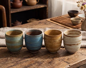 May include: Four ceramic cups with a rustic, textured finish. Each cup has a unique color: teal, blue, golden brown, and off-white. The cups are cylindrical and sit on a wooden table, suggesting a tea or coffee set.