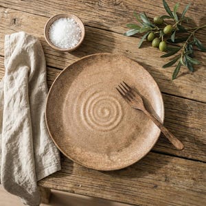 May include: A brown ceramic plate with a concentric circle design, a wooden fork, a small bowl of salt, a linen napkin, and olive branch on a wooden table. The plate has a textured surface. The scene has a natural, earthy aesthetic.