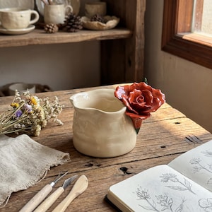 May include: A cream-coloured ceramic jug with a red rose detail. The rose is fixed to the side of the jug. The jug is on a wooden surface. Other ceramic items are on a shelf in the background.