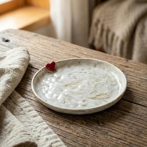 May include: A handmade, off-white ceramic plate with a small, red heart accent. The plate has a textured surface and a slightly irregular shape. It sits on a rustic wooden table next to a linen cloth. The plate is suitable for serving food.