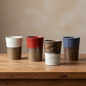 May include: Four ceramic tumblers with a two-tone design. The top halves are white, red, brown, and blue, while the bottom halves are a dark brown. The tumblers are displayed on a wooden surface.