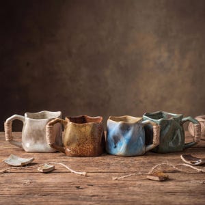 May include: Four handcrafted ceramic mugs in varying colors: off-white, brown, blue, and green. Each mug has a unique textured surface and a square shape with a wavy rim. The handles are wrapped with twine. The mugs are arranged on a wooden surface.