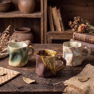 May include: Three handcrafted ceramic mugs with unique, textured designs. One is green, another is brown and tan, and the third is white and tan. Each mug has a handle and sits on a wooden table, with a rustic background.