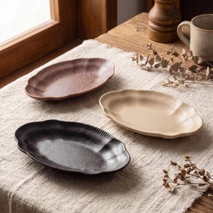 May include: Three ceramic oval serving platters in shades of brown, beige, and black. Each platter has a scalloped edge and a textured surface. The platters are arranged on a neutral-colored linen tablecloth.