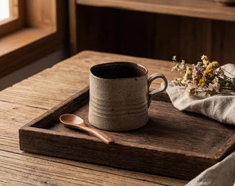 Puede incluir: Una taza de cerámica con interior marrón y exterior marrón claro con líneas horizontales se encuentra sobre una bandeja de madera. Una pequeña cuchara de madera descansa junto a la taza. La bandeja está sobre una mesa de madera, con flores secas y un paño de lino.