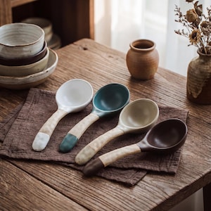 May include: A set of four ceramic spoons with rounded bowls and textured, light-coloured handles. Each spoon features a distinct coloured tip in white, teal, beige, and dark brown, matching the interior colour of its respective bowl.