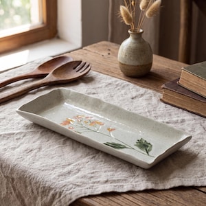May include: A rectangular ceramic serving tray with an off-white speckled finish and a hand-painted floral design featuring orange blossoms and green leaves. The tray has slightly raised, uneven edges and a smooth, glossy glaze surface.