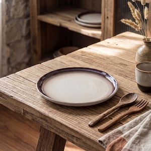 May include: A cream-coloured ceramic plate with a dark brown rim rests on a rustic wooden table. A wooden spoon and fork are placed beside the plate. A small ceramic cup and a vase with dried plants are also on the table.