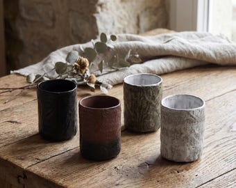 May include: Four cylindrical ceramic cups with textured, uneven surfaces. One cup is solid black, one is a gradient of reddish-brown to black, one is olive green with a mottled finish, and one is white with a visible grid-like fabric texture.
