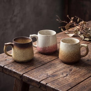 Może przedstawiać: Trzy ceramiczne kubki z nakrapianymi wykończeniami w odcieniach brązu, beżu i bieli. Każdy kubek ma zaokrąglony korpus i zakrzywiony uchwyt. Kubki są ułożone na rustykalnej drewnianej powierzchni.