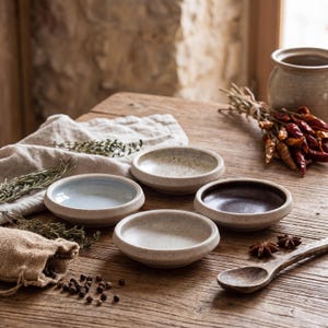 May include: A collection of small, handcrafted ceramic bowls in various neutral tones, including beige, light blue, and brown, arranged on a rustic wooden table. The scene includes a wooden spoon, spices, and a small burlap bag.