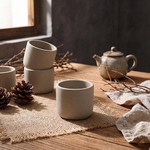 Puede incluir: Un conjunto de tazas de té de cerámica de tonos neutros y una tetera dispuestas sobre una mesa de madera. Las tazas son cilíndricas con un acabado moteado. Piñas y ramitas complementan la estética natural.