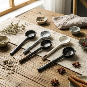 Könnte beinhalten: Eine Sammlung von fünf Keramik-Löffeln mit langen Griffen, jeweils mit einem einzigartigen gesprenkelten oder einfarbigen Design. Die Löffel sind auf einem Holztisch mit kleinen Schalen und Gewürzen angeordnet, was eine rustikale und elegante Ästhetik schafft.