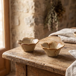 Puede incluir: Dos cuencos pequeños de cerámica beige con diseño de pétalos de flor sobre una mesa de madera rústica. Los cuencos tienen una superficie texturizada y un borde marrón oscuro. El fondo presenta una pared de piedra y plantas secas.