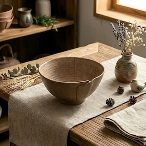 Può includere: Una grande ciotola in ceramica marrone strutturata è appoggiata su un runner da tavolo in lino beige. La ciotola ha un'ampia apertura e una forma leggermente irregolare. Il tavolo è in legno e ha un aspetto rustico. Un piccolo vaso con fiori secchi è anche sul tavolo.