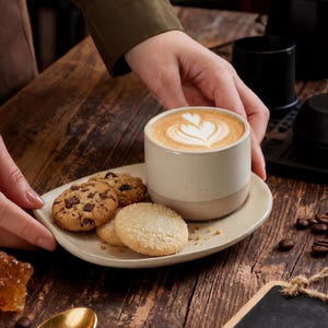 Hands placing a handmade ceramic cup on an oval stoneware saucer with latte art and cookies
