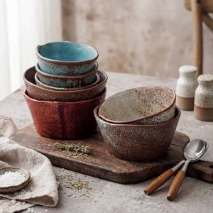 Puede incluir: Un conjunto de cuencos de cerámica rústica en tonos rojos, marrones y verde azulado, con superficies texturizadas. Los cuencos están colocados sobre una tabla de madera, junto con saleros y pimenteros, cucharas con mangos de madera y hierbas.
