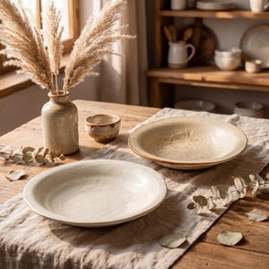 Peut inclure: Une table de salle à manger rustique avec deux bols en céramique crème, une petite tasse assortie et un vase avec de l'herbe de la pampa séchée. La table est recouverte d'un tissu en lin, créant une atmosphère chaleureuse.