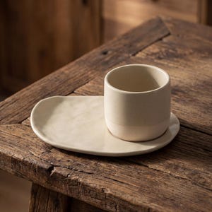 Minimalist handmade ceramic cup and oval saucer set on rustic wooden table, speckled stoneware finish
