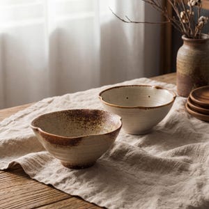 Pode incluir: Duas tigelas de cerâmica com uma aparência rústica e artesanal. As tigelas são de cor branco-sujo com detalhes salpicados castanhos nas bordas e superfícies externas. Elas estão sobre uma toalha de linho bege em uma mesa de madeira, sugerindo uma estética natural.