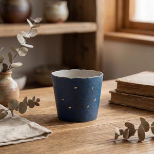 Puede incluir: Una taza de cerámica azul marino con borde festoneado y motas doradas. La taza está sobre una mesa de madera, con un pequeño jarrón y hojas secas en el fondo. La taza mide aproximadamente 10 cm de alto.