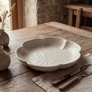 Op de afbeelding: Een grote, gebroken witte, bloemblad-vormige serveerschaal met een textuuroppervlak. De schaal staat op een houten tafel naast een linnen servet, mes en vork. Decoratieve vazen met gedroogde planten staan op de achtergrond.