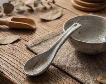 May include: A ceramic soup spoon with a speckled, light gray finish, resting on a linen cloth. A matching bowl and wooden scoop are also visible, suggesting a rustic, minimalist aesthetic. The spoon's design features a small hole at the end of the handle.