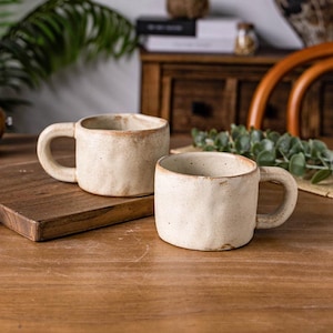 May include: Two off-white ceramic mugs with handles. The mugs have a rustic, textured appearance with a light brown rim. They are placed on a wooden surface, suggesting a cozy, handcrafted aesthetic. The mugs are ideal for coffee or tea.