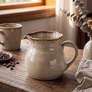 May include: A ceramic pitcher with a glossy cream-colored glaze and a scalloped rim featuring a dark brown drip-style accent. The pitcher has a rounded body and a curved handle, showcasing a smooth, reflective surface and a rustic aesthetic.