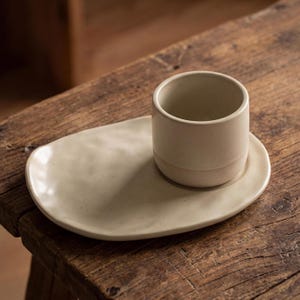 Minimalist handmade ceramic cup and oval saucer set on rustic wooden table, speckled stoneware finish