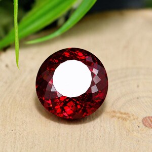 Pode incluir: Uma pedra preciosa facetada, redonda, vermelho escuro com um centro branco brilhante. A gema está sobre uma superfície de madeira castanha clara, com folhagem verde ao fundo. Esta gema pode ser usada em joias.