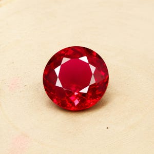 Può includere: Una gemma di rubino rotonda e sfaccettata, di colore rosso intenso. La gemma è posta su uno sfondo beige chiaro, evidenziando la sua taglio brillante e le sue sfaccettature riflettenti. Il rubino ha una forma circolare e sembra essere una gemma di alta qualità.