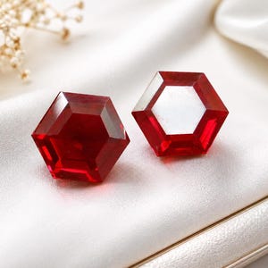 May include: A pair of vibrant red, hexagonal-shaped faceted glass or acrylic gems. Each piece features a flat top surface with beveled edges that create a geometric, multi-sided appearance, reflecting light across the deep crimson surfaces.
