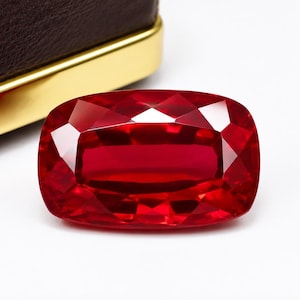 Puede incluir: Una gema roja intensa, tallada en forma rectangular y facetada. La gema pulida refleja la luz, mostrando sus facetas internas. La piedra preciosa se encuentra sobre un fondo blanco, con parte de un estuche marrón y dorado visible.