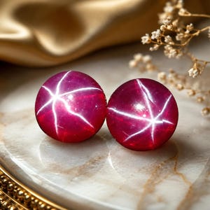 Könnte beinhalten: Zwei runde, tief magentafarbene Cabochon-Edelsteine, die jeweils ein ausgeprägtes sechsstrahliges Sternmuster auf der Oberfläche aufweisen. Die Steine haben eine glatte, polierte Oberfläche und eine leuchtende, gesättigte rot-violette Farbe.