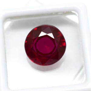 May include: A faceted, round-cut ruby gemstone. The gem is a deep, rich red color with multiple facets that reflect light. The ruby is set against a white background, highlighting its color and clarity. The gemstone is likely for jewelry.