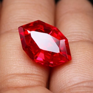 Op de afbeelding: Een gefacetteerde, levendige rode edelsteen met een unieke langwerpige vorm. De edelsteen heeft meerdere reflecterende oppervlakken, die licht vangen en een briljante uitstraling creëren. De steen wordt in een hand gehouden, wat de grootte en kleur benadrukt.